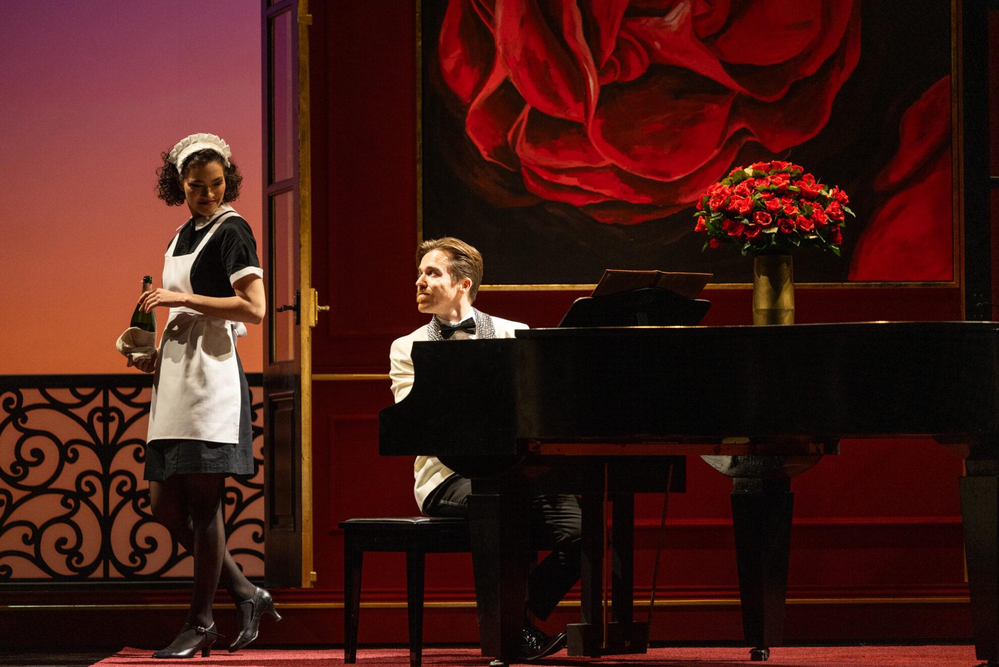Doug Kelly behind a piano, looking at Nina Korbe standing